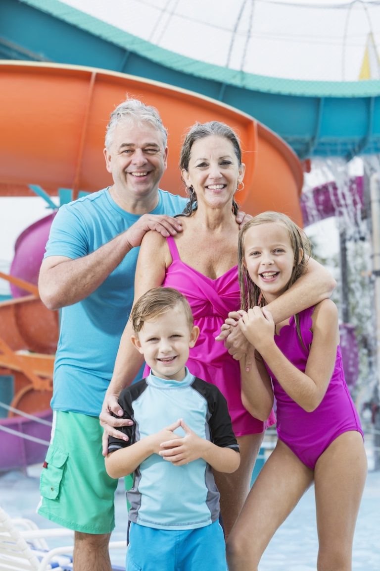 Two children with their grandparents having fun at a water park, standing in front of a giant water slide, smiling at the camera. The girl is 10 years old and her brother is 6.